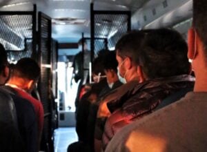 People wearing masks standing in a crowded bus aisle, metal shelves along the sides, dim lighting, looking forward.