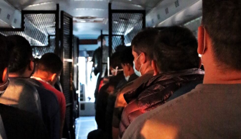 People wearing masks standing in a crowded bus aisle, metal shelves along the sides, dim lighting, looking forward.