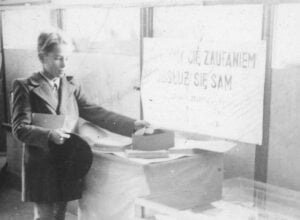 Black-and-white photo of a man in a suit sitting at a desk with papers, facing left; a blurred sign with faded text is behind him.