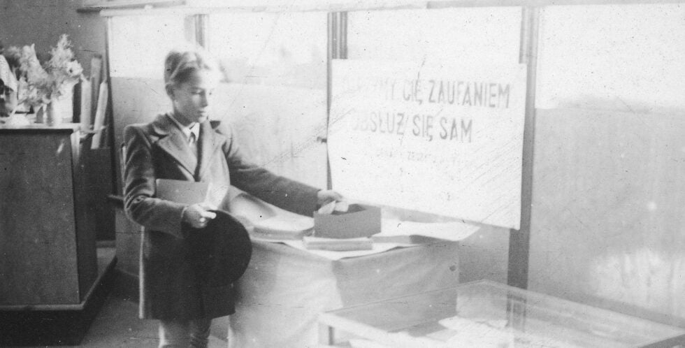 Black-and-white photo of a man in a suit sitting at a desk with papers, facing left; a blurred sign with faded text is behind him.