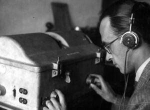 Person wearing over-ear headphones leans in to adjust knobs on a vintage control panel.