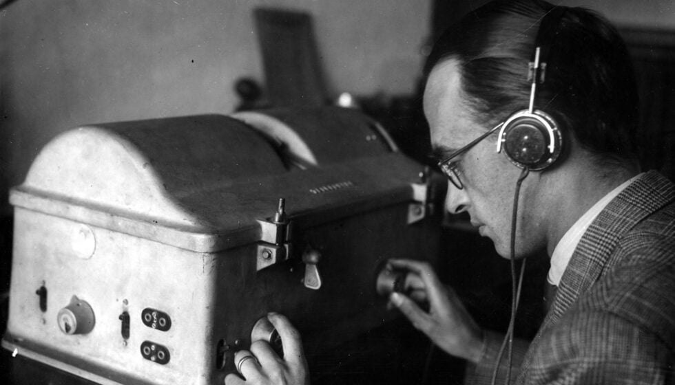 Person wearing over-ear headphones leans in to adjust knobs on a vintage control panel.