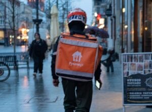 Back view of a delivery rider in a helmet with an orange delivery backpack on a rainy city street
