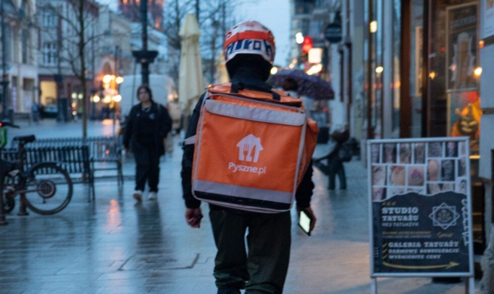 Back view of a delivery rider in a helmet with an orange delivery backpack on a rainy city street