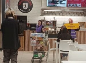 Customer wearing a black coat stands in front of a kebab shop counter in a mall food court, with staff in purple vests behind the counter and a shopper cart nearby.