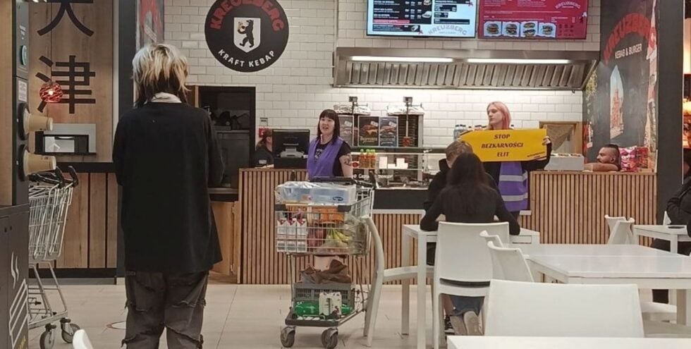 Customer wearing a black coat stands in front of a kebab shop counter in a mall food court, with staff in purple vests behind the counter and a shopper cart nearby.