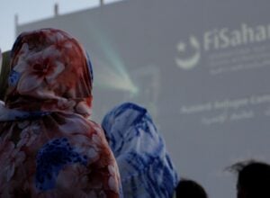 People wearing colorful floral hoodies seen from behind at an outdoor event with a large blurred screen in the background.