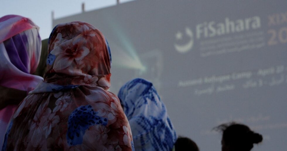 People wearing colorful floral hoodies seen from behind at an outdoor event with a large blurred screen in the background.