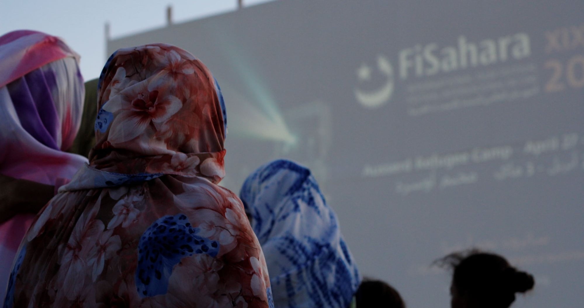 People wearing colorful floral hoodies seen from behind at an outdoor event with a large blurred screen in the background.