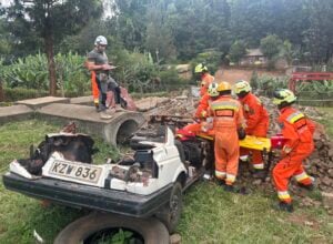 Emergency responders in orange uniforms and helmets assess a collapsed area with a wrecked car and debris; a supervisor notes on a clipboard nearby (license plate KZW 836).