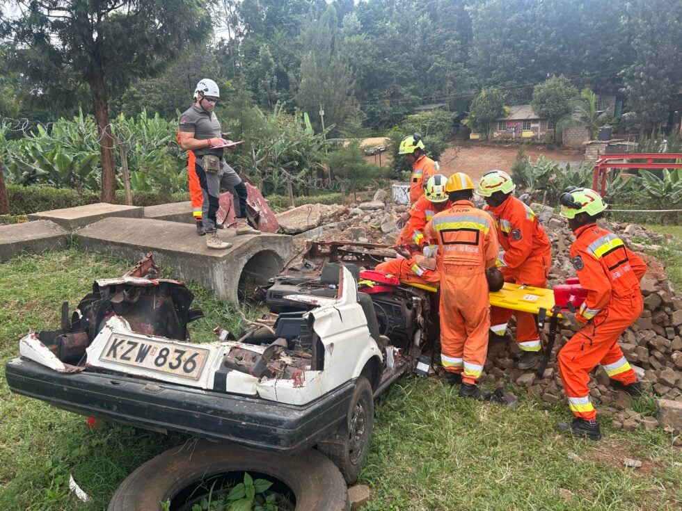 Emergency responders in orange uniforms and helmets assess a collapsed area with a wrecked car and debris; a supervisor notes on a clipboard nearby (license plate KZW 836).