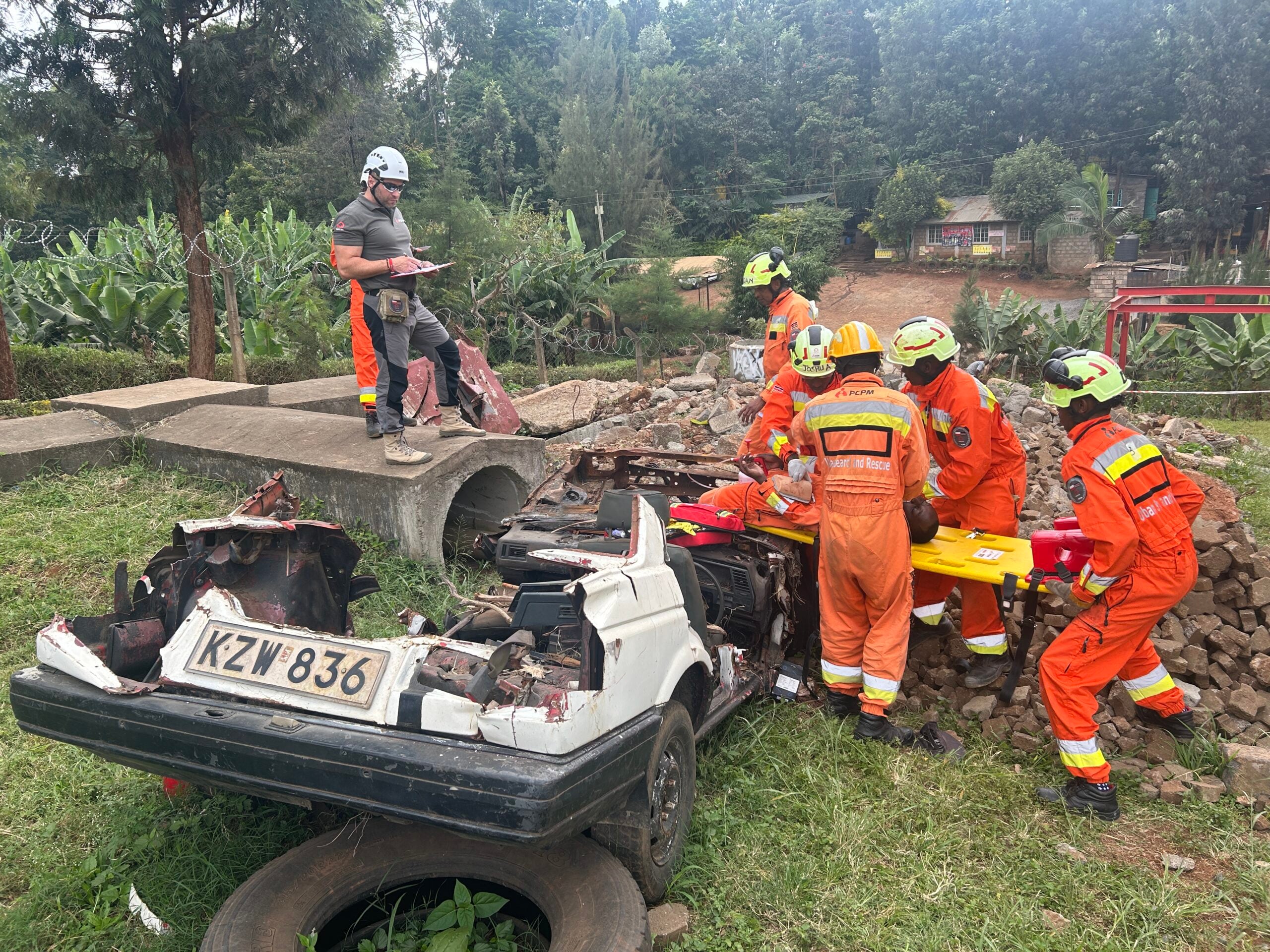 Emergency responders in orange uniforms and helmets assess a collapsed area with a wrecked car and debris; a supervisor notes on a clipboard nearby (license plate KZW 836).