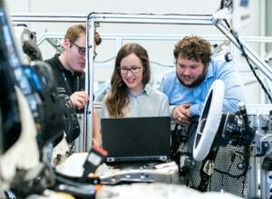 Three engineers gathered around a laptop inside a high-tech manufacturing setup, inspecting equipment near metal framing and cables.