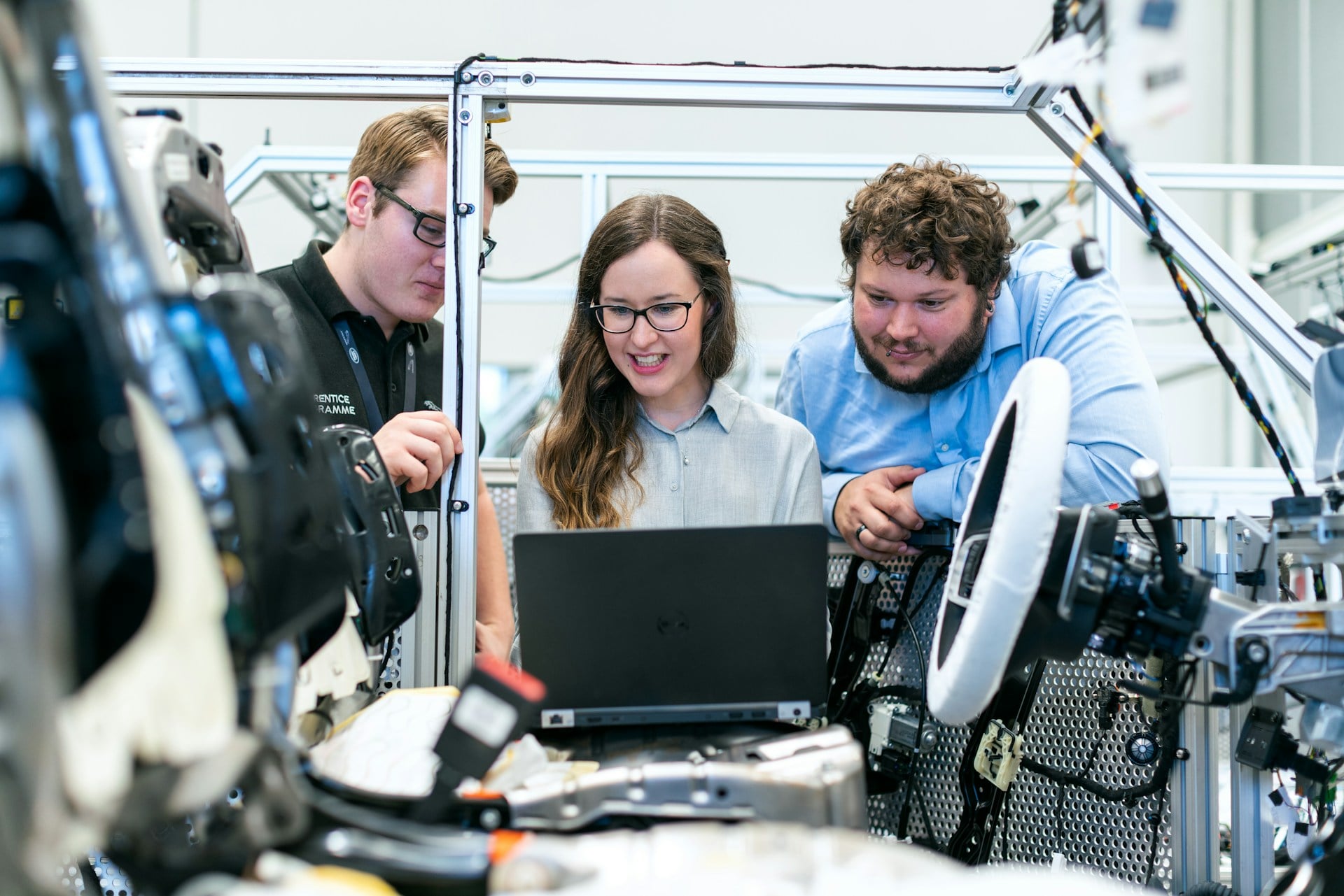 Three engineers gathered around a laptop inside a high-tech manufacturing setup, inspecting equipment near metal framing and cables.