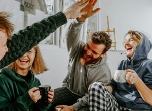 Four friends in a bright room high-five each other, holding mugs and smiling. An upbeat, casual hangout scene.