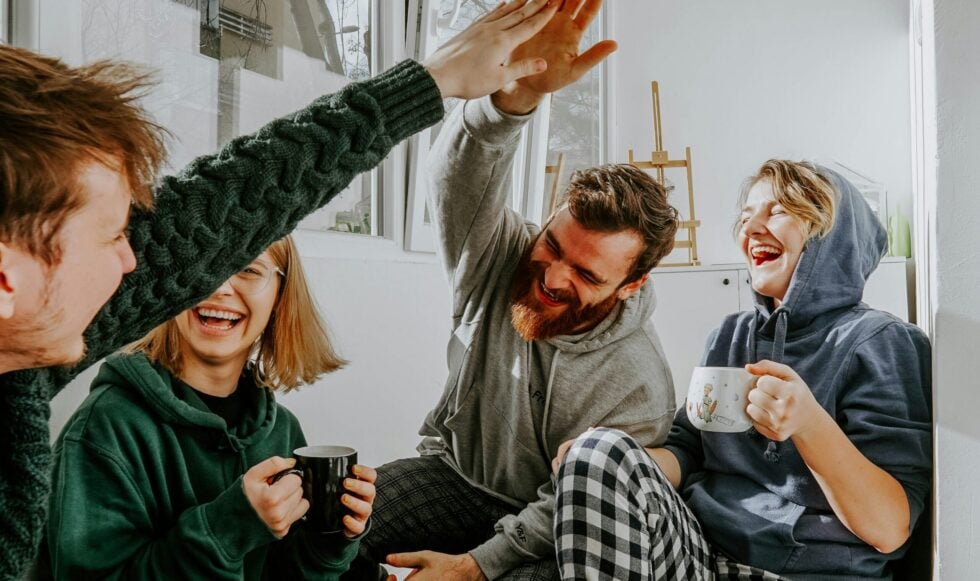 Four friends in a bright room high-five each other, holding mugs and smiling. An upbeat, casual hangout scene.