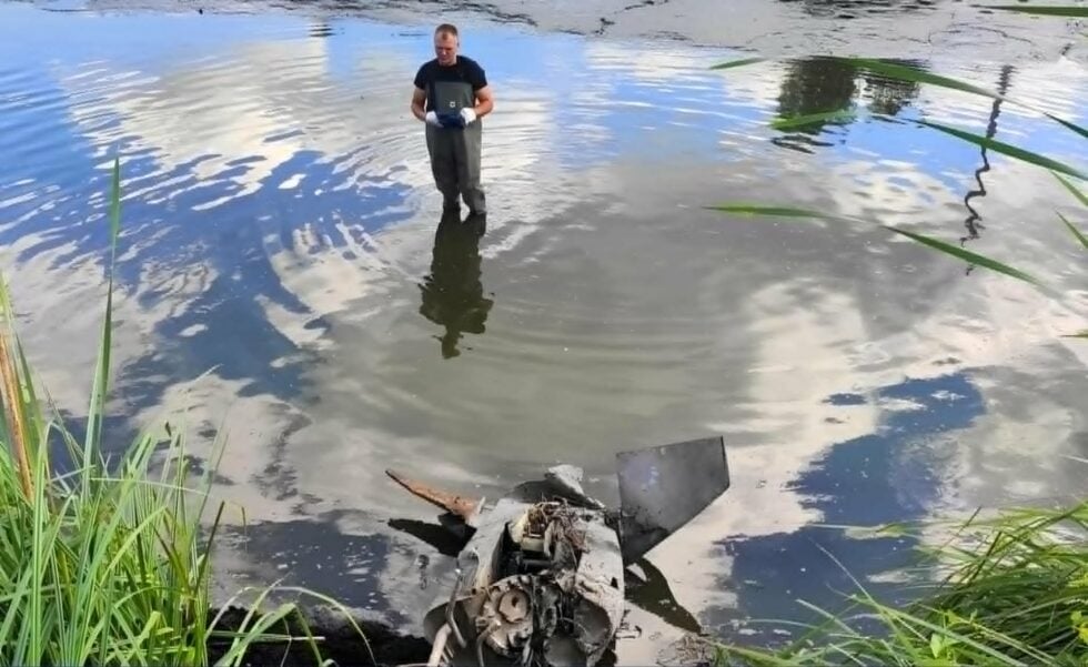 Man standing in a shallow pond wearing chest-high waders and gloves, examining the water amid reeds and debris.