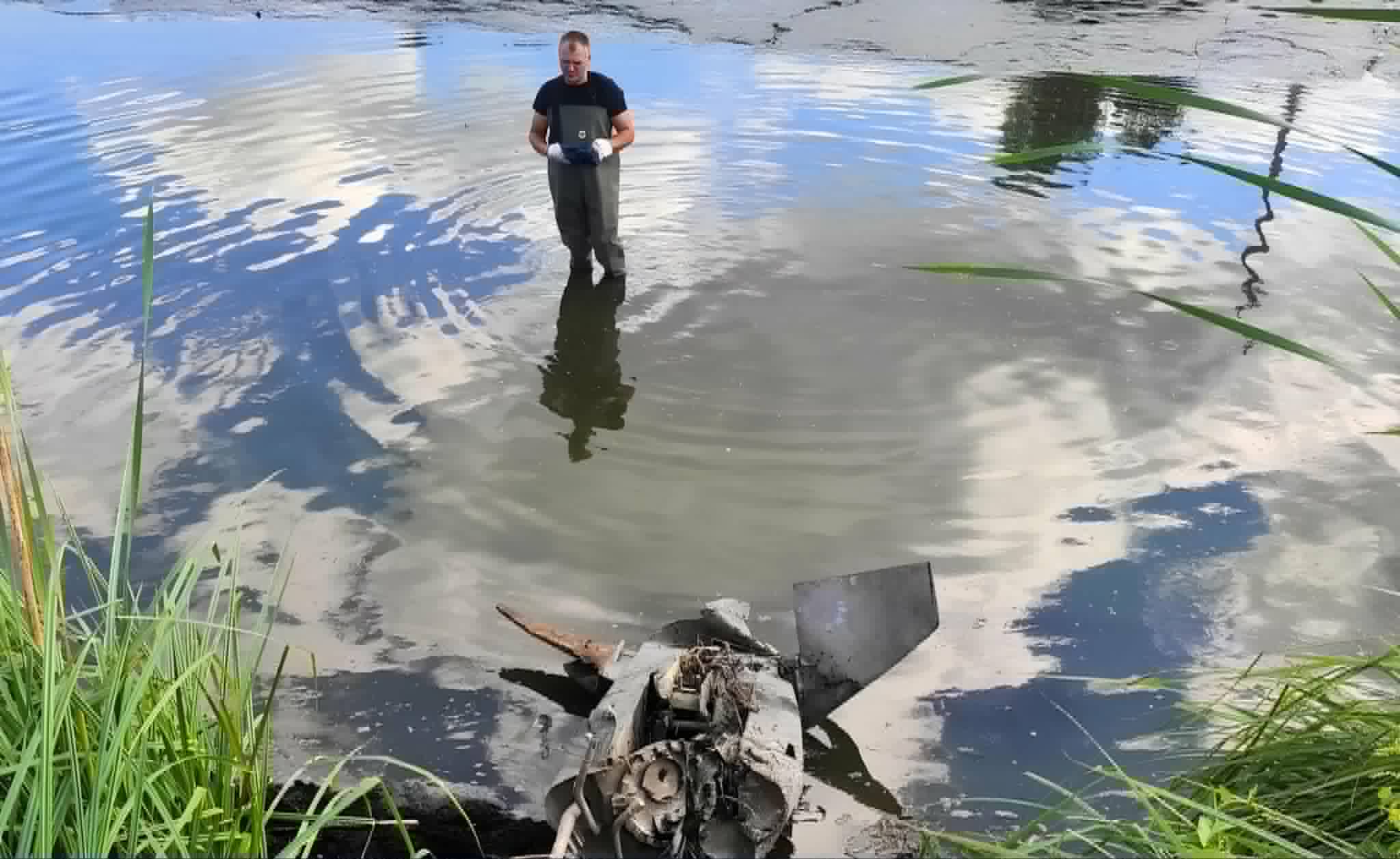 Man standing in a shallow pond wearing chest-high waders and gloves, examining the water amid reeds and debris.