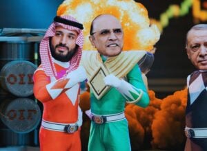 Three costumed performers stand together in front of a bright explosion backdrop, with oil drums stacked on the left.