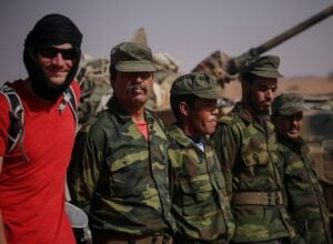Group of men in camouflage uniforms standing beside a military vehicle in a desert setting, one man in a red shirt with a black headwrap.