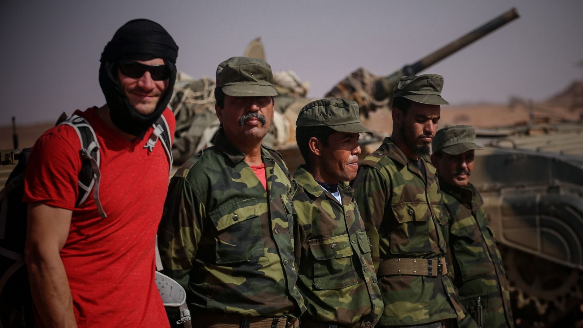 Group of men in camouflage uniforms standing beside a military vehicle in a desert setting, one man in a red shirt with a black headwrap.