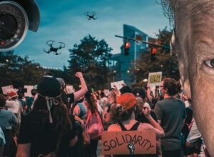 Protestujący tłum widziany od tyłu, widoczna tabliczka z napisem "Solidarity", na niebie w lewym górnym rogu kamaera inwigilijąca i dwa drony, po prawej stronie połowa twarzy Trumpa