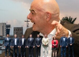 A bald man in a suit points toward the camera at a harbor, with a line of formally dressed people standing along the water behind him.