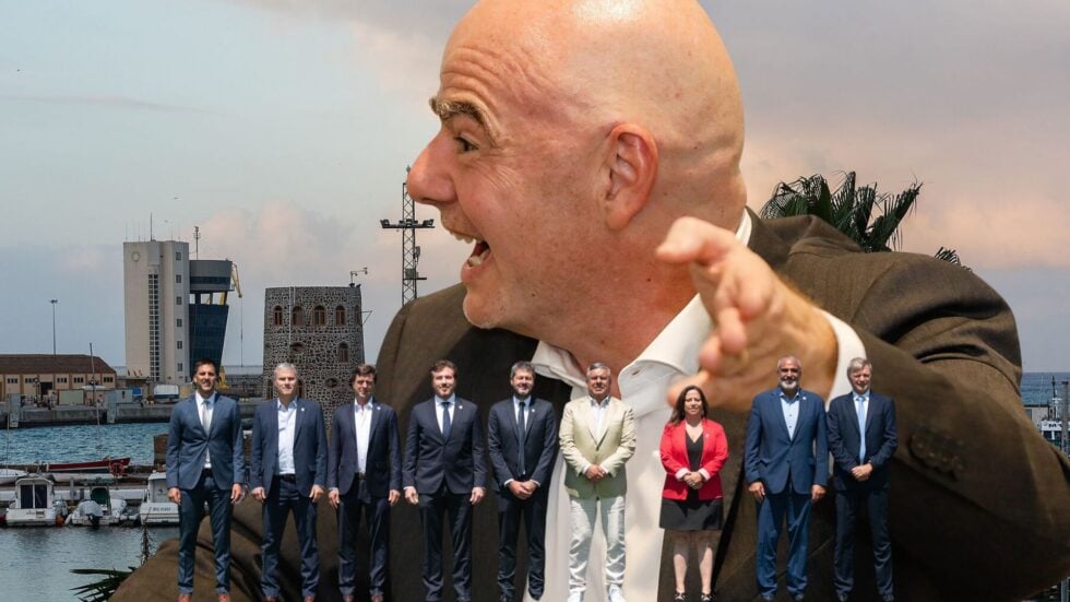 A bald man in a suit points toward the camera at a harbor, with a line of formally dressed people standing along the water behind him.