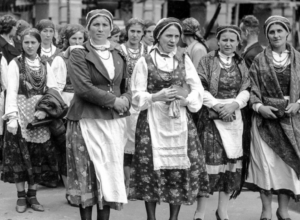 Women in traditional folk dresses stand in a line outdoors, some holding shawls and bags.