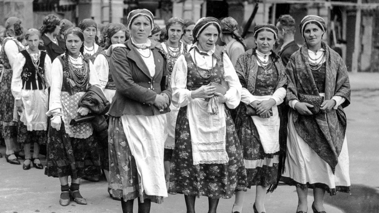 Women in traditional folk dresses stand in a line outdoors, some holding shawls and bags.