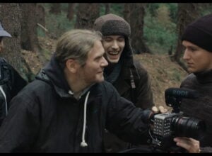 A film crew in a forest gathers around a camera, sharing a laugh while filming outdoors.
