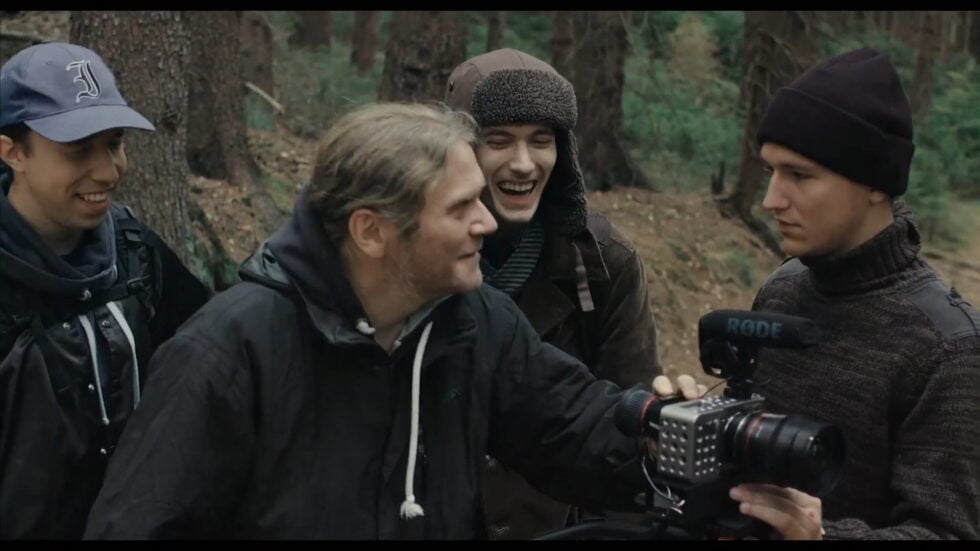 A film crew in a forest gathers around a camera, sharing a laugh while filming outdoors.