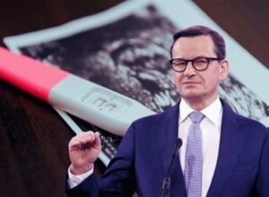 Businessman in a suit and glasses speaking at a podium, with a large positive pregnancy test prop in the foreground and a photo in the background.
