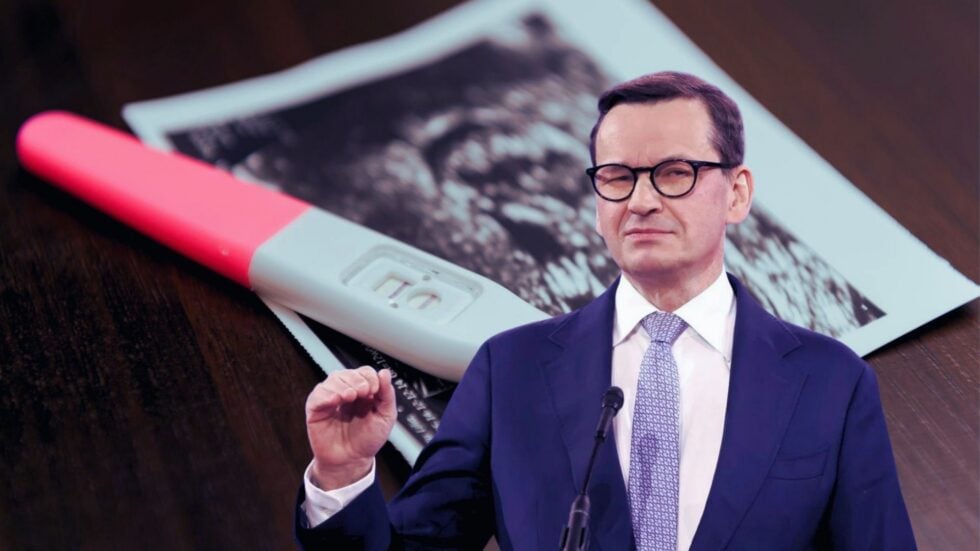 Businessman in a suit and glasses speaking at a podium, with a large positive pregnancy test prop in the foreground and a photo in the background.