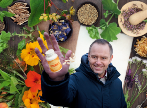 Smiling man in a dark jacket holds a vitamin bottle toward the camera, with herbs and spices spread on a wooden table behind.