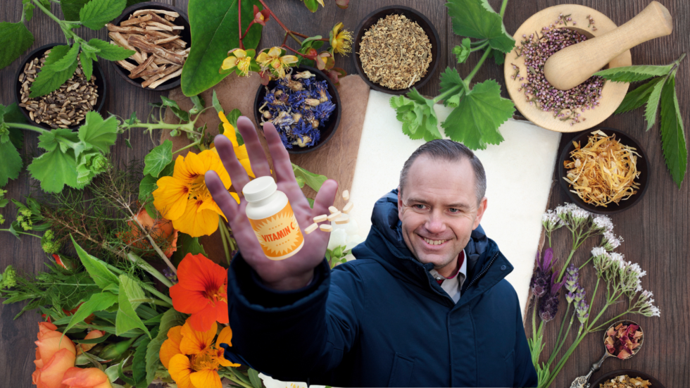 Smiling man in a dark jacket holds a vitamin bottle toward the camera, with herbs and spices spread on a wooden table behind.