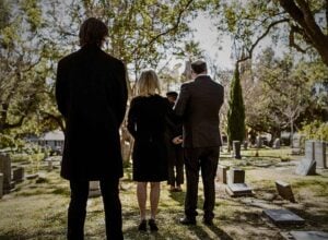Back view of formally dressed mourners standing in a sunlit cemetery among headstones and trees.