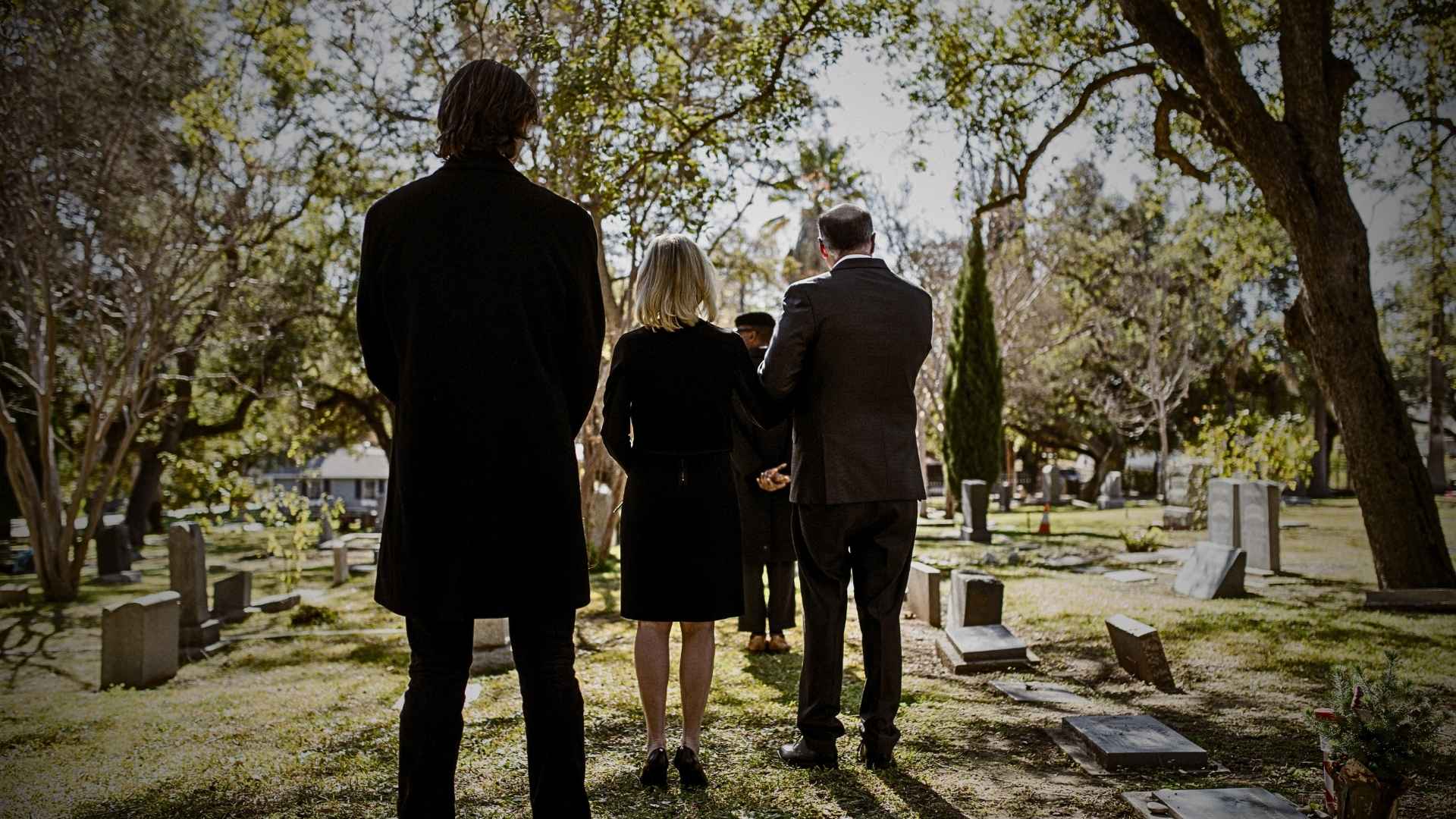 Back view of formally dressed mourners standing in a sunlit cemetery among headstones and trees.
