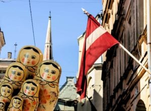 Cluster of colorful Matryoshka dolls stacked in a narrow city street with a red-and-white flag overhead against a blue sky.