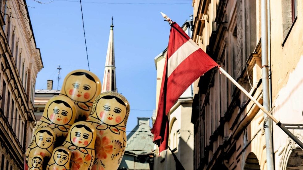 Cluster of colorful Matryoshka dolls stacked in a narrow city street with a red-and-white flag overhead against a blue sky.