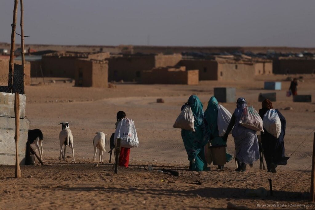 Women in traditional robes carrying cloth sacks walk through a sandy desert village with goats nearby.