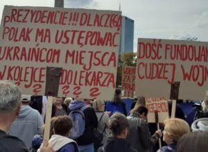 Crowd of protesters holding large cardboard signs with red handwritten Polish slogans outdoors; one sign reads 'DOŚĆ FUNDOWANIA CUDZEJ WOJNY' in bold letters.