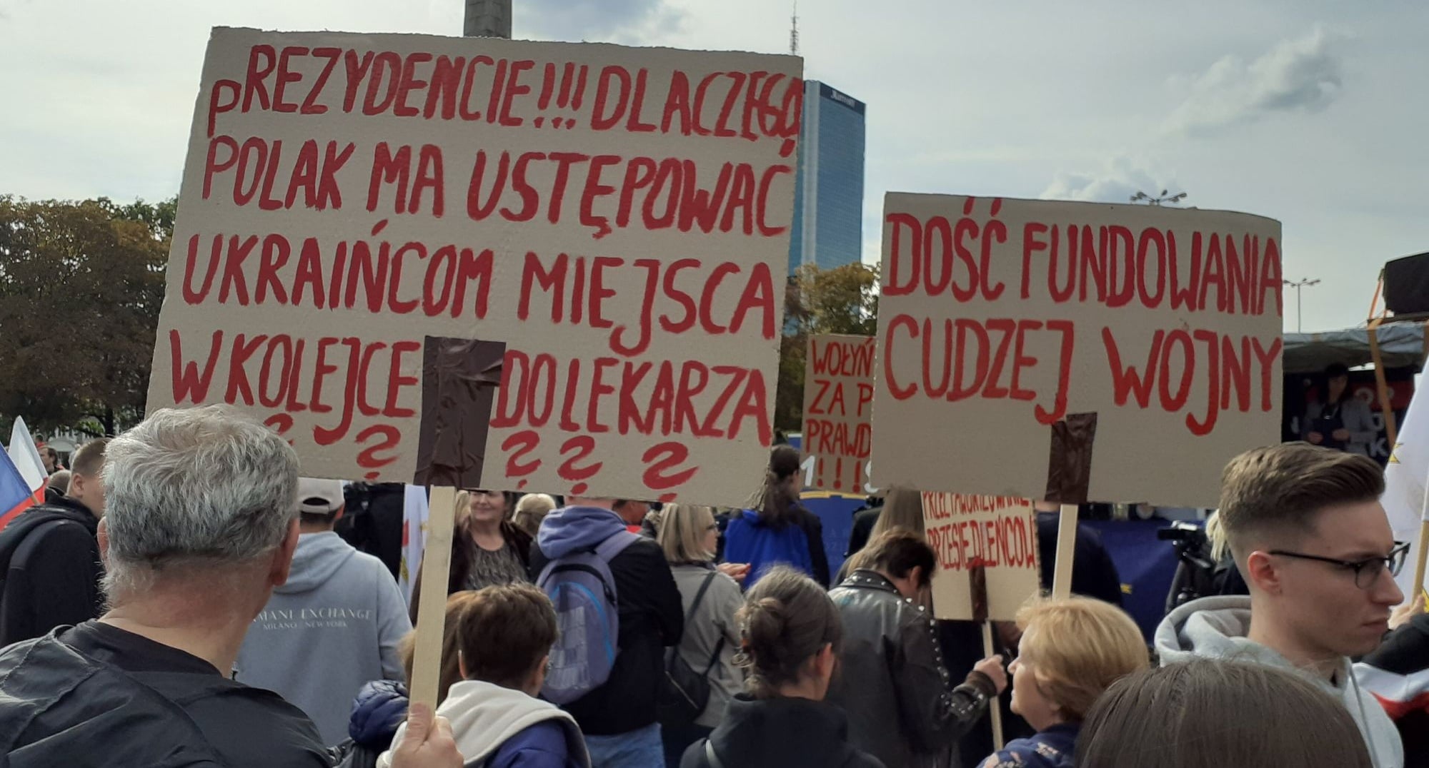 Crowd of protesters holding large cardboard signs with red handwritten Polish slogans outdoors; one sign reads 'DOŚĆ FUNDOWANIA CUDZEJ WOJNY' in bold letters.