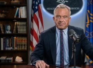 Man in a navy suit speaks into a microphone at a desk, with the U.S. flag and a blue banner behind him in a book-lined office.