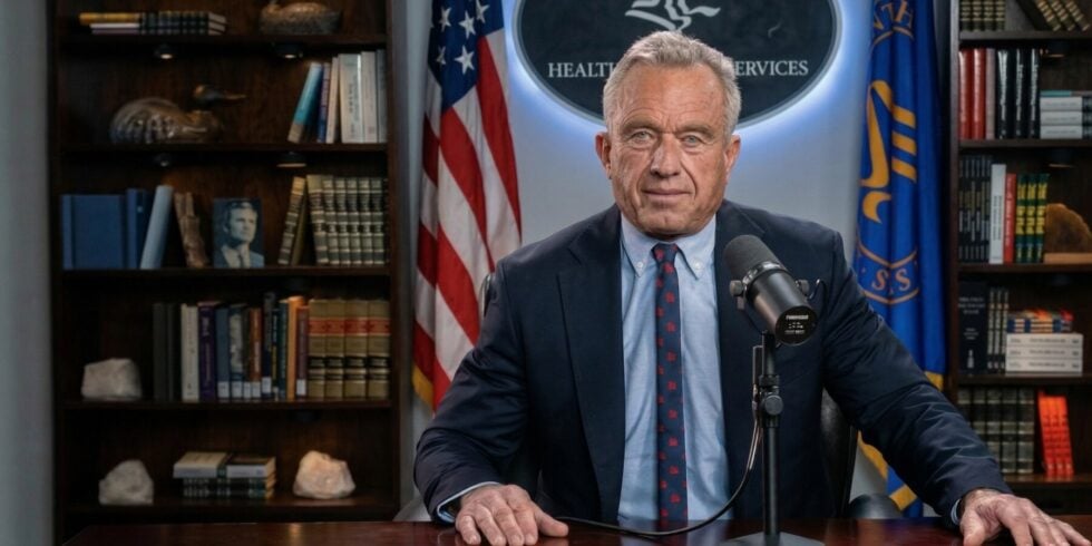 Man in a navy suit speaks into a microphone at a desk, with the U.S. flag and a blue banner behind him in a book-lined office.