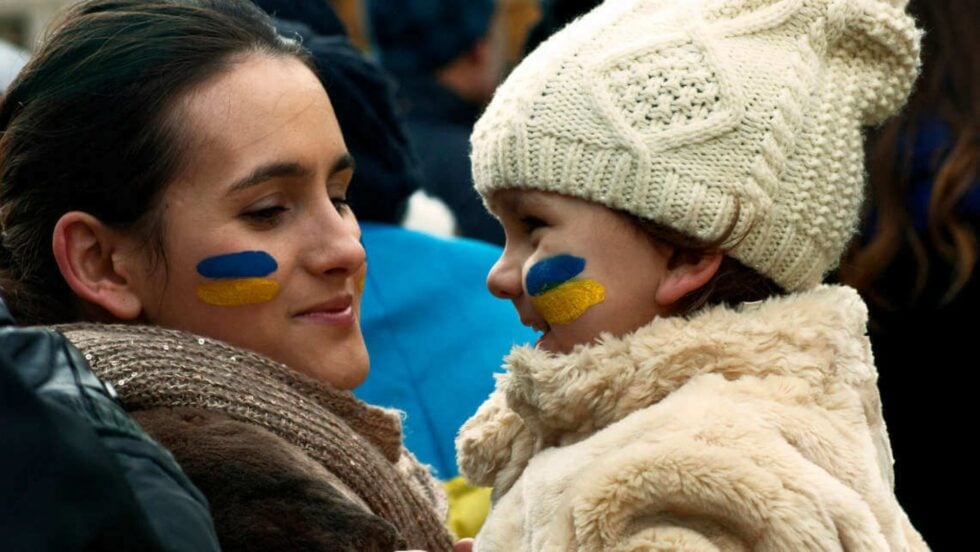 Two people facing each other with blue and yellow face paint, wearing warm coats and a cream knit hat on one side.
