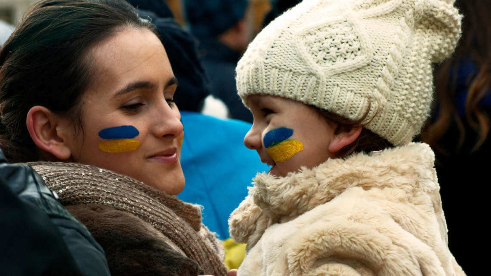 Two people facing each other with blue and yellow face paint, wearing warm coats and a cream knit hat on one side.