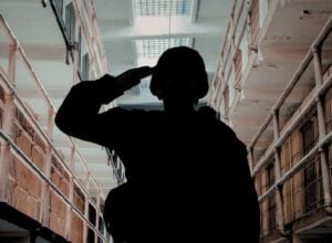 Silhouette of a person saluting in a prison corridor with barred cell doors on both sides perhaps a hallway.