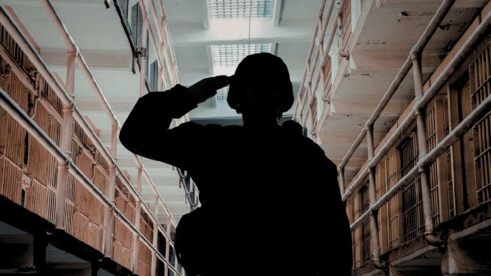 Silhouette of a person saluting in a prison corridor with barred cell doors on both sides perhaps a hallway.