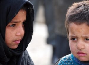 Two children stand side by side, the girl in a black scarf and the boy in a blue sweater, both looking somber.